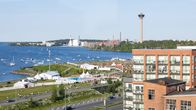 Aurinkoinen kesäpäivä Tampereen Santalahdessa, jossa ihmiset nauttivat festivaalitunnelmasta ja järvimaisemasta. Taustalla näkyy kaupungin siluetti ja lukuisat veneet vesillä.