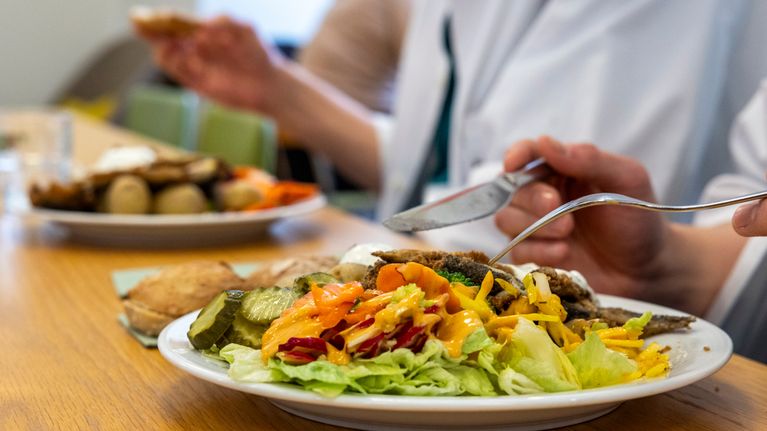 Två händer med kniv och gaffel i vardera hand i färd med att äta mat från en tallrik med bland annat sallad och saltgurka.