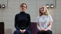 vå personer sitter bredvid varandra på en bänk inomhus. Den ena bär en svart polo och byxor, den andra har på sig en vit t-shirt och mörka byxor.