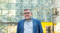 Juhani Sand hymyilee. Hän seisoo keskellä Taysin pääsisäänkäynnin aulatilaa. Hänellä on päällään sininen puvuntakki. 