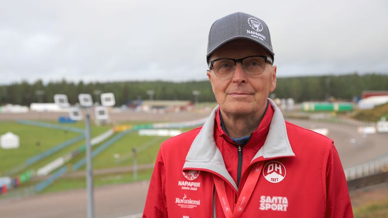 Napapiiri-Jukolan kilpailunjohtaja Martti Anttila punaisessa takissa ja harmaassa lakissa, taustalla Mäntyvaaran ravirataa.