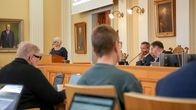 En kvinna står vid en talarstol i en sal med flera porträtt och stora vita pelare, personer sitter vid datorer.