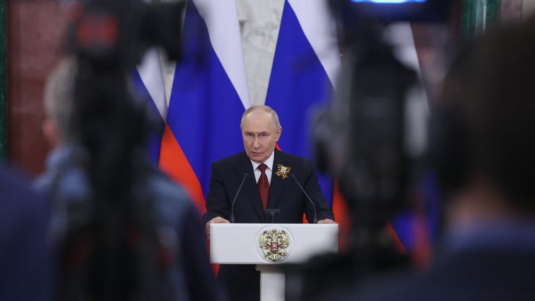 Vladimir Putin står vid ett talarpodium framför en rad ryska flaggor. Han syns mellan fotografer och journalister.