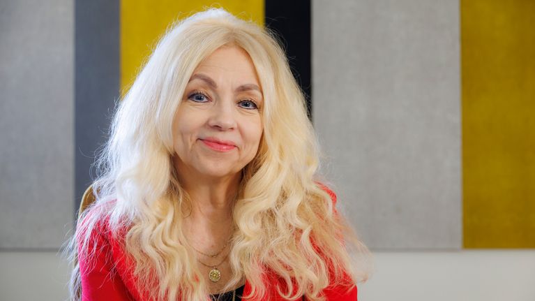 Marita Husso i en röd tröja ler och tittar in i kameran. Hon sitter på en stol och i bakgrunden syns en randig vägg.