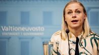 A blonde woman in a beige-and-green top speaks at a press conference with a government logo behind her.