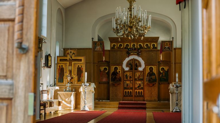 The interior of an ornate Orthodox church features a grand chandelier, icon-adorned wooden altar, and rich red carpeted aisle leading to intricately designed iconostasis panels.