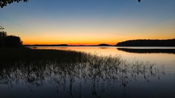 Järvimaisema auringonlaskun aikaan; taivas oranssin ja sinisen sävyissä, järven pinnalla heijastuksia.