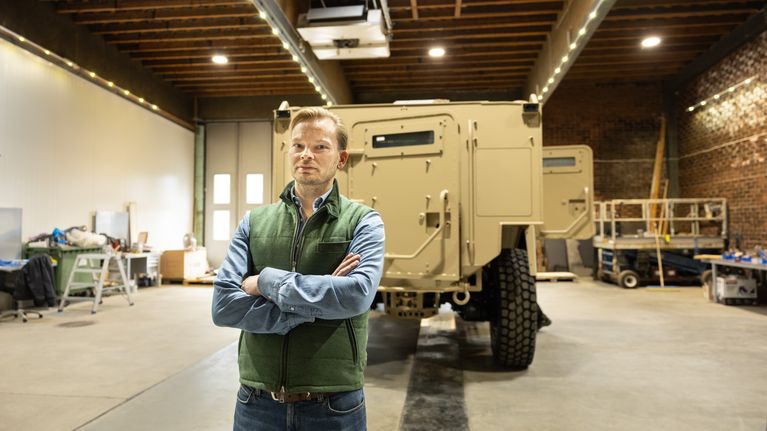 En person står med korsade armar framför ett stort militärfordon i en verkstadslokal.