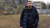 Henkilö seisoo ulkona kevättalvisessa maisemassa kivimuurin ja rakennusten edustalla. Hänellä on yllään tumma talvitakki ja musta huppari. Maassa on vähän ruskeaa ruohoa näkyvissä, mikä viittaa siihen, että kevät on alkamassa.
