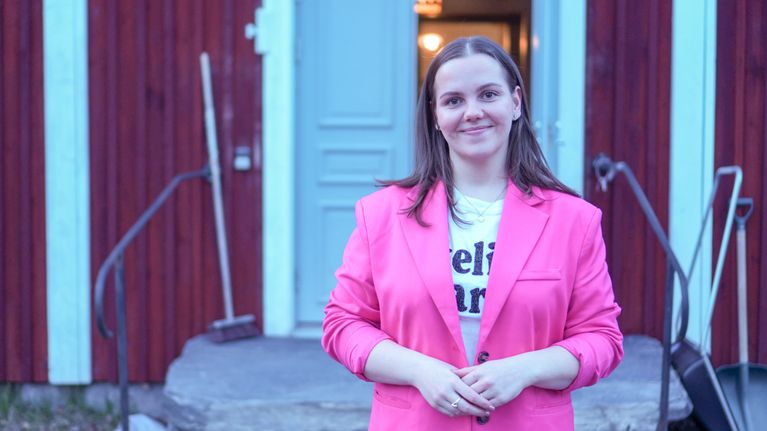Pinkkiin takkiin pukeutunut Maria Tokou hymyilee kameralle punaisen puurakennuksen edessä.
