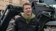 En person med jacka står framför en grön traktor och staplade träskivor.