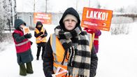 Lakkovahti Amelia Kiiltomäki katsoo kameraan. Taustalla muita lakkovahteja kylttien kanssa talvisessa maisemassa.