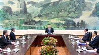 Xi Jinping istuu pöydän päässä. Hänen takanaan on kiinalaista vuorimaisemaa esittävä kuva.