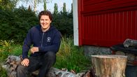 En person sitter på en stor trädstam intill en röd byggnad, med gräs och buskar i bakgrunden.