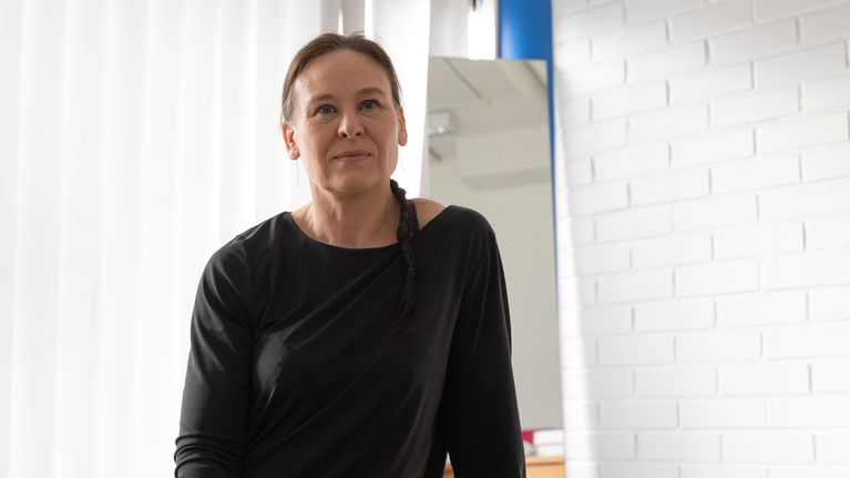 Tässä kuvassa on henkilö, joka istuu huoneessa. Hänellä on musta paita, ja hänen hiuksensa ovat letillä.