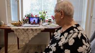 Ikääntynyt nainen istuu nojatuolissa ja katsoo pöydällä olevaa tablettitietokonetta, jossa on auki etävideopuhelu.