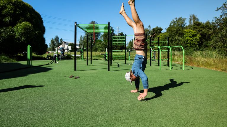 Lippispäinen mies seisoo käsillään ulkokuntosalin nurmimatolla.