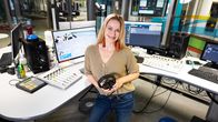 Henkilö seisoo modernissa radiostudiossa, pitäen kuulokkeita käsissään. Studiota ympäröivät näytöt ja äänentoistolaitteet,.