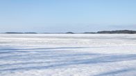 A frozen lake with snow on it, a bluish sky above and treelined shores and islands on the distant horizon.  
