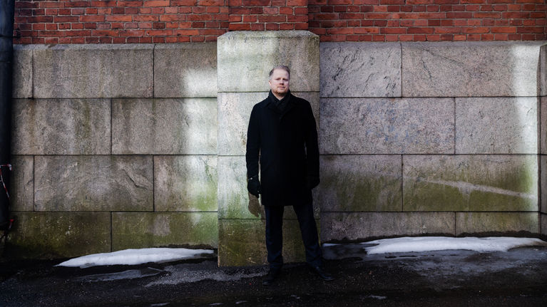 A man wearing a dark jacket and pants standing in front of a grey stone and red brick building.