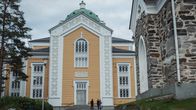 Kolme henkilöä seisoo valtavan kokoisen, keltaisen puukirkon edustalla.