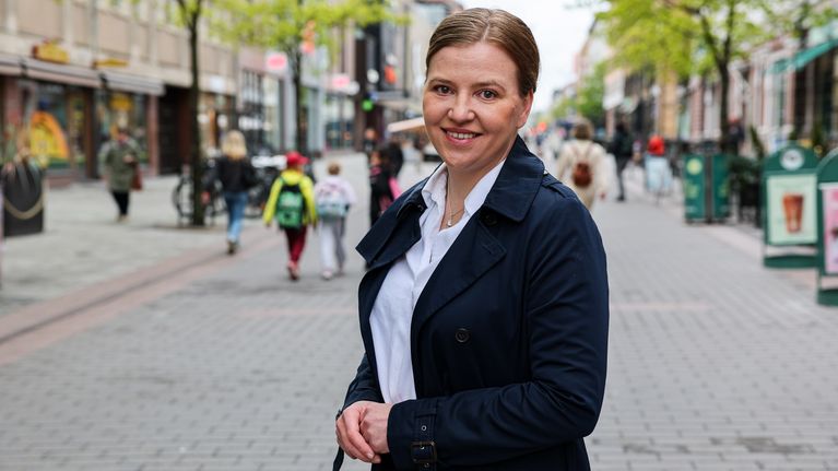 En person i mörk kappa står på en stadsgata med träd och människor i bakgrunden.