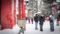 People wearing winter clothes standing among red buildings and snow-covered trees.