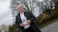 Henkilö pukeutuneena valkoiseen paitaan ja tummaan takkiin ulkona. Hän näyttää kameralle kädessään olevaa kirjaa sunny car centerin tarinasta.