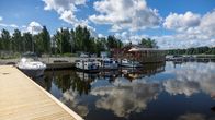 Venesatama, jossa useita veneitä laiturissa kirkkaassa säässä; taustalla metsää ja rakennuksia.