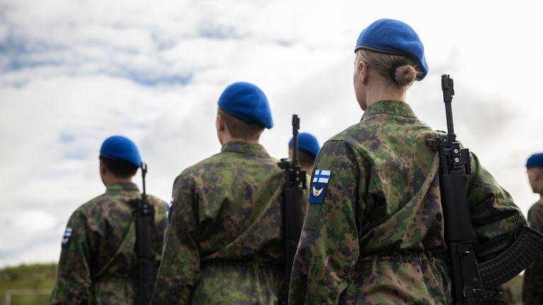 Three Air Force conscripts standing in a line, seen from behind, with the one on the right with her blonde hair in a bun under a blue beret.