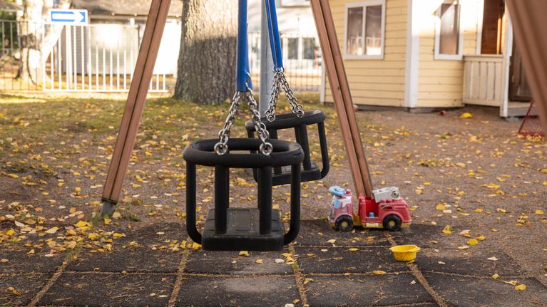 Två tomma gungor på en liten lekplats, i bakgrunden syns en urblekt leksaksbrandbil.