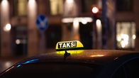 Keltainen taksikyltti tumman auton katolla, taustalla kaupunkimiljöötä illalla.