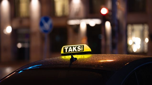 Keltainen taksikyltti tumman auton katolla, taustalla kaupunkimiljöötä illalla.