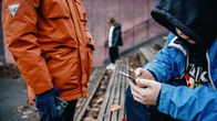 Two children wearing autumn weather jackets, one looking at a smartphone, with a child in the background, just out of focus.