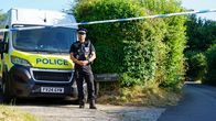 Poliisi seisoo korkean pensaan vieressä, takanaan poliisiauto.