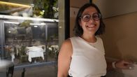 A woman wearing glasses stands by a window in a dimly lit restaurant, smiling at the camera.