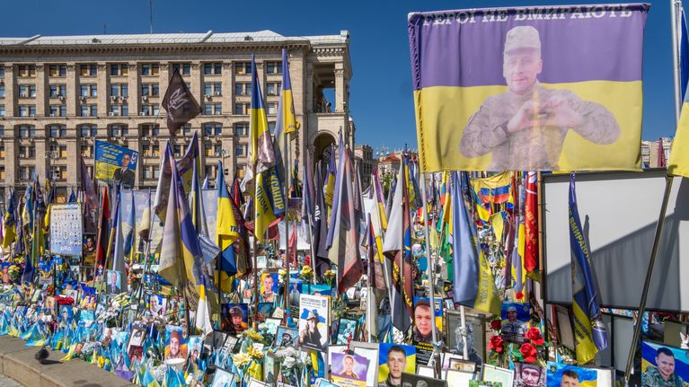 Porträttfoton, ukrainska flaggor och blommor är uppställda utomhus vid en minnesplats framför en byggnad. På en stor banderoll syns en person och text på ukrainska högst upp.
