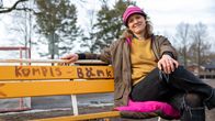 En kvinna som sitter på en bänk som är målad med texten "kompis-bänk". Personen är klädd i en gul tröja, en brun jacka, rosa mössa och svarta jeans. 