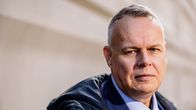 Mies seisoo pukeutuneena siniseen pukuun ja solmioon, yllään tumma päällystakki, ulkona rakennuksen edustalla.