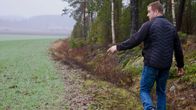 En man med jeans och svart jacka går längs en skogskant och pekar mot ett öppet fält.