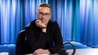 A man wearing glasses and a black zipper jacket sits with a half-smile in a radio studio with a headset on the table.