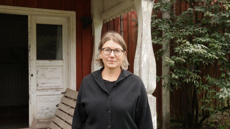 En person med glasögon och svart tröja står framför en röd träbyggnad med vit dörr.