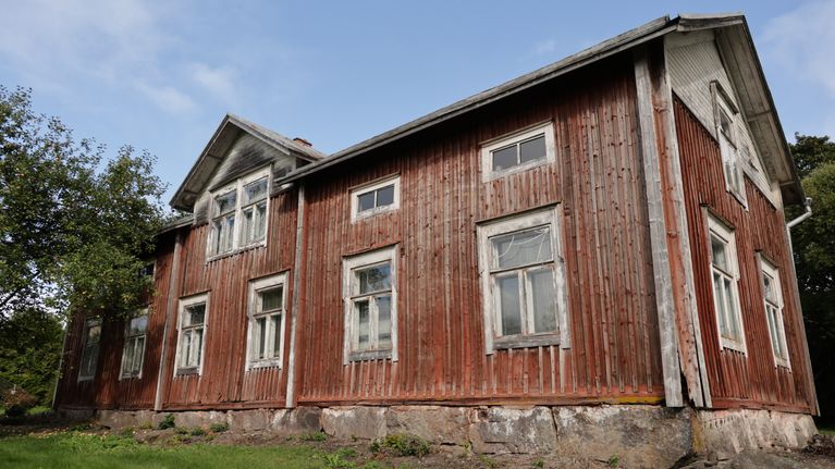 Röd trähusbyggnad med vita fönsterramar och stengrund, omgiven av gräs och träd under blå himmel.