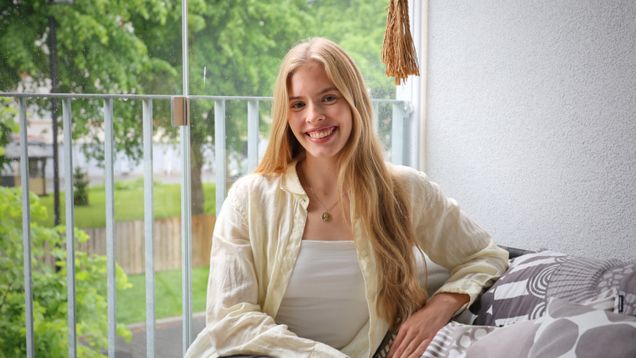 En person med långt blont hår sitter på en balkongsoffa intill ett räcke, med träd och gräs utomhus i bakgrunden.
