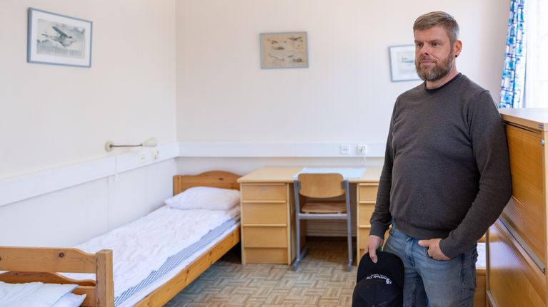 Mathias Blomqvist står i ett enkelt möblerat sovrum med en säng, ett skrivbord och en stol.