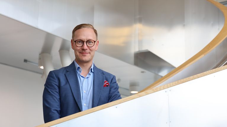 En man med blå kavaj, blå skjorta och glasögon står i en trappa och tittar in i kameran. 