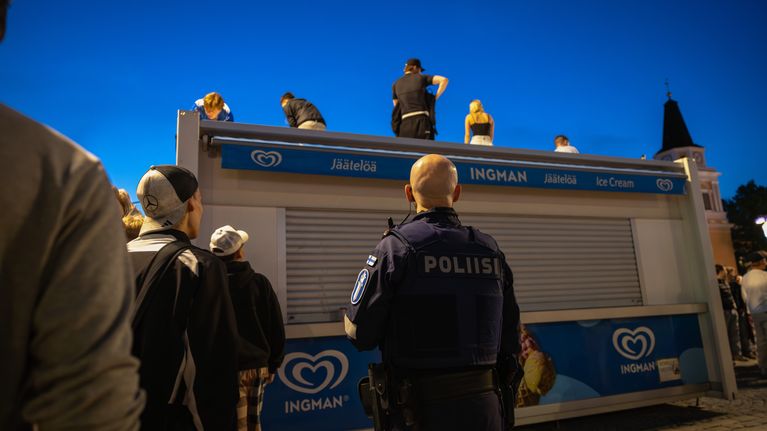 Poliisi seisomassa selin kameraan, katsoen nuoria, jotka ovat kiivenneet Keskustorin jäätelökioskin päälle.