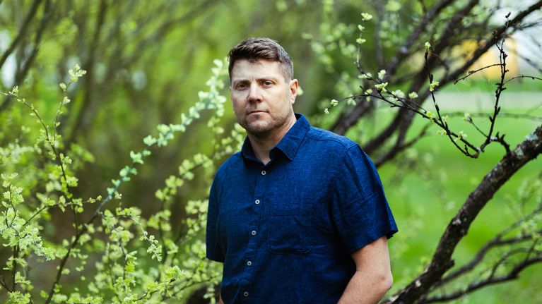 Thomas Ramstedt, en av Svenska Yles Sommarpratare 2026, står utomhus bland gröna trädgrenar.