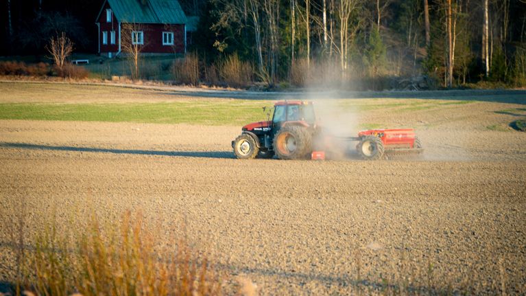 A red tractor pulls a small trailer in a dusty field with a small red wooden house and trees in the distance.