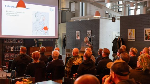 En person håller en presentation inför en sittande publik; en karta och texten "Esiintymisalueella" syns på en projicerad skärm.